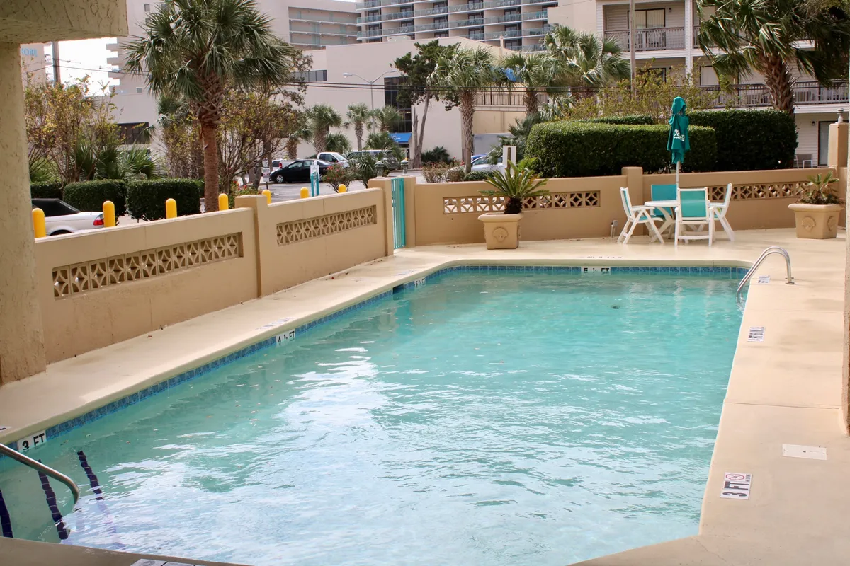 A lively exterior shot of a resort-style swimming pool surrounded by lounge chairs and palm trees. The pool area is sunlit, inviting guests to swim, sunbathe, or gather with friends and family—emphasizing the leisure amenities available just steps from the condo.