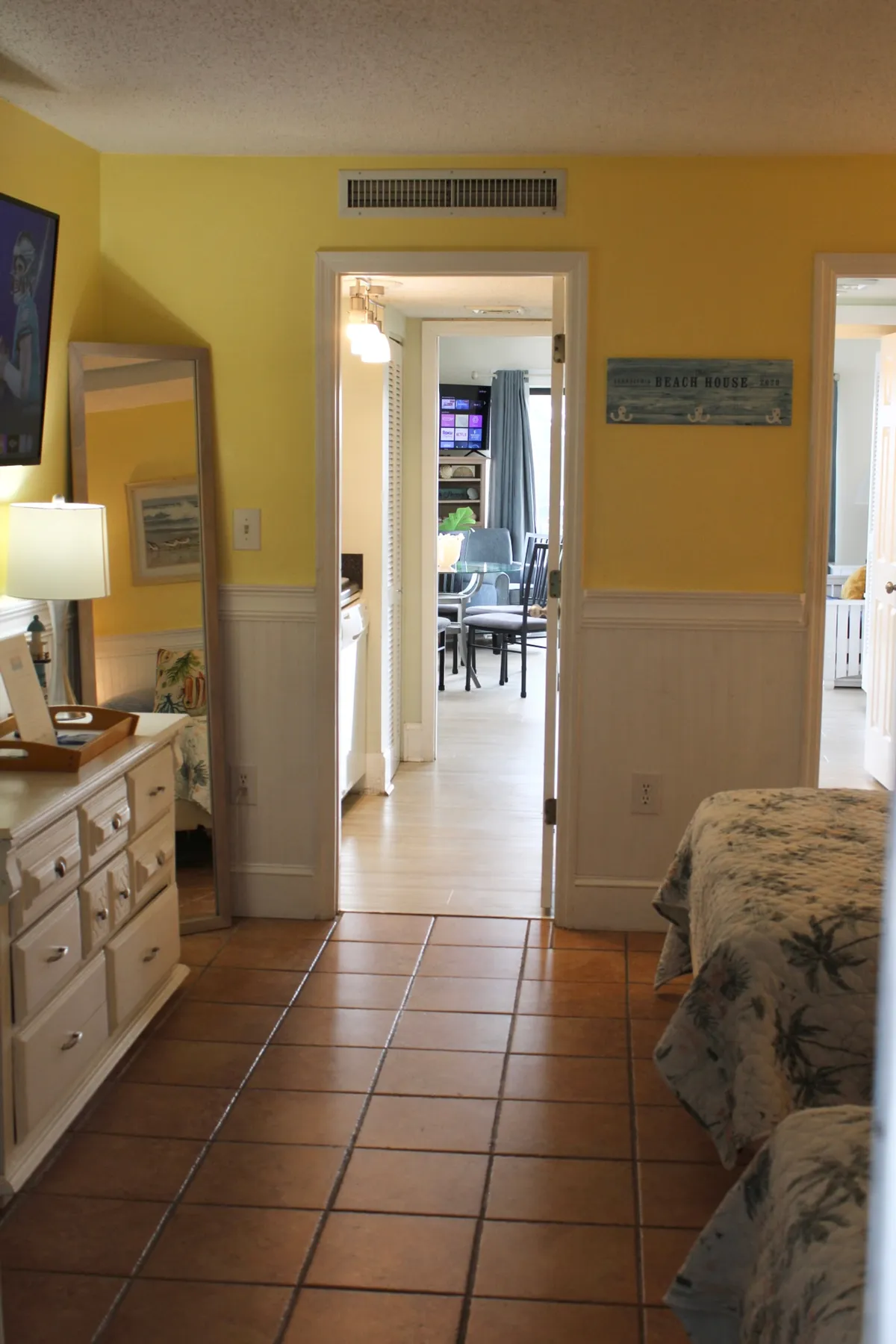 This image features two beds with floral bedding, a wall-mounted TV, a dresser with decorative items, and a mirror that expands the sense of space. A doorway leads to another room, hinting at additional sleeping or living areas. The atmosphere is casual and welcoming.