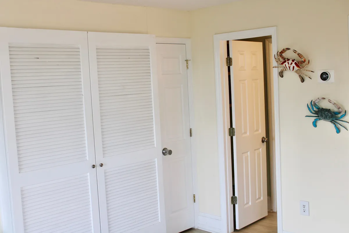 A light-filled, inviting room with white louvered closet doors, playful crab wall art, and a glimpse into an adjacent space. The decor hints at a laid-back coastal lifestyle, ready for guests to personalize with their own beach finds or store their gear after a day by the ocean.