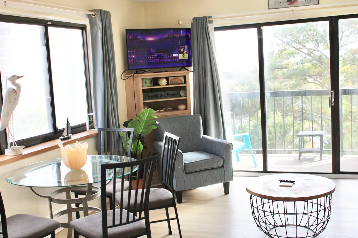 A different angle of the living area, this scene features a glass dining table set near a cozy armchair and TV, with decorative items on the shelf and a glimpse of the balcony. The setup invites relaxation and shared experiences, all within an open, airy environment.