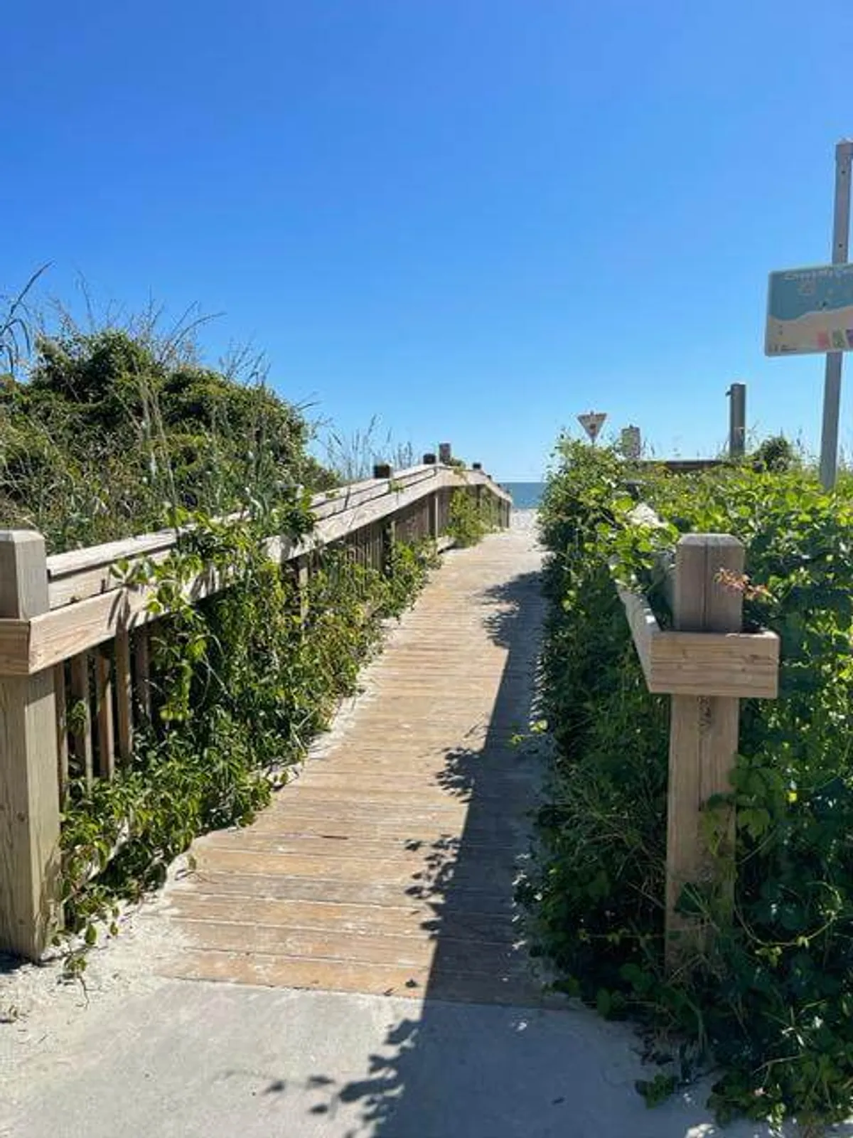 A tranquil exterior view showing a wooden pathway leading through lush greenery toward the distant ocean. The scenery evokes a sense of peaceful anticipation, with the promise of beach adventures and natural beauty just ahead.
