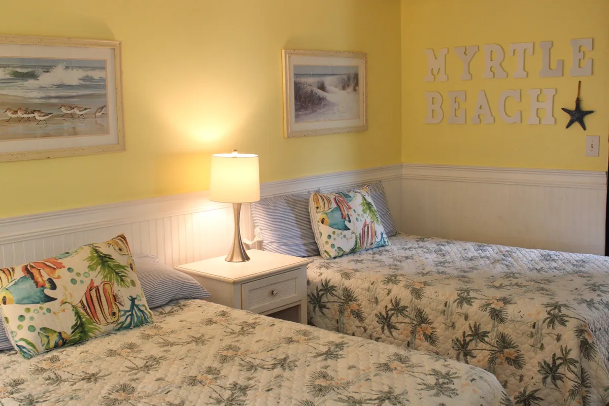 The image showcases a bright, inviting bedroom with two neatly made beds adorned with decorative pillows, a cozy lamp on a bedside table, and cheerful coastal decor including a 'Myrtle Beach' sign and beach-themed artwork. Sunlight filters through a nearby window, creating a warm and welcoming atmosphere perfect for relaxation.
