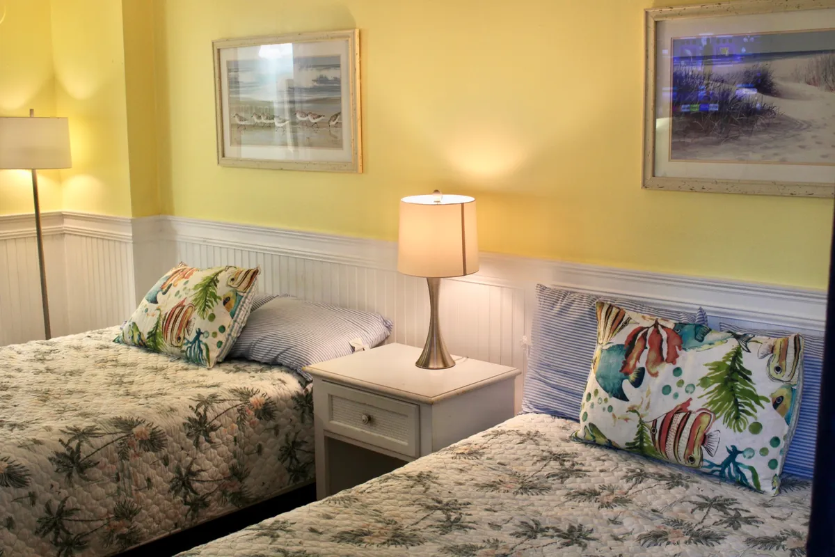 Showing two twin beds with decorative pillows, a table lamp, and framed artwork, this bedroom is styled for comfort and relaxation. The mood is cozy and encourages socializing or relaxing mornings and evenings.