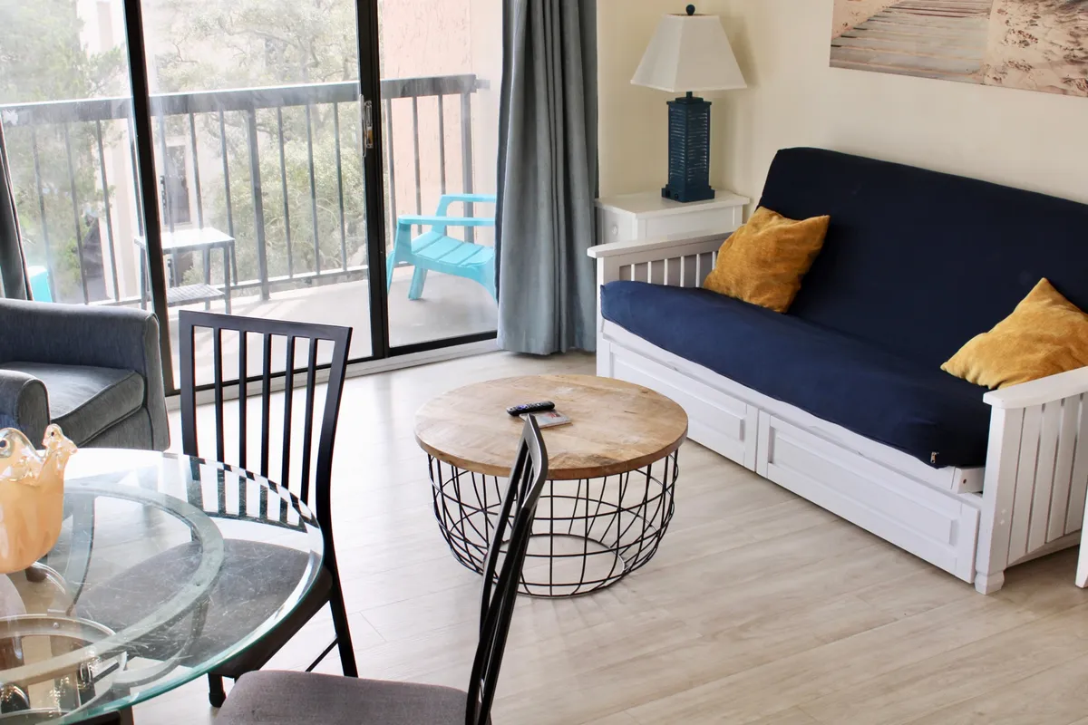 This image displays a plush sofa with gold pillows facing a glass dining table, complemented by a stylish wireframe coffee table and blue chairs visible on the balcony. The space is sunlit and inviting, ideal for both relaxation and group activities.