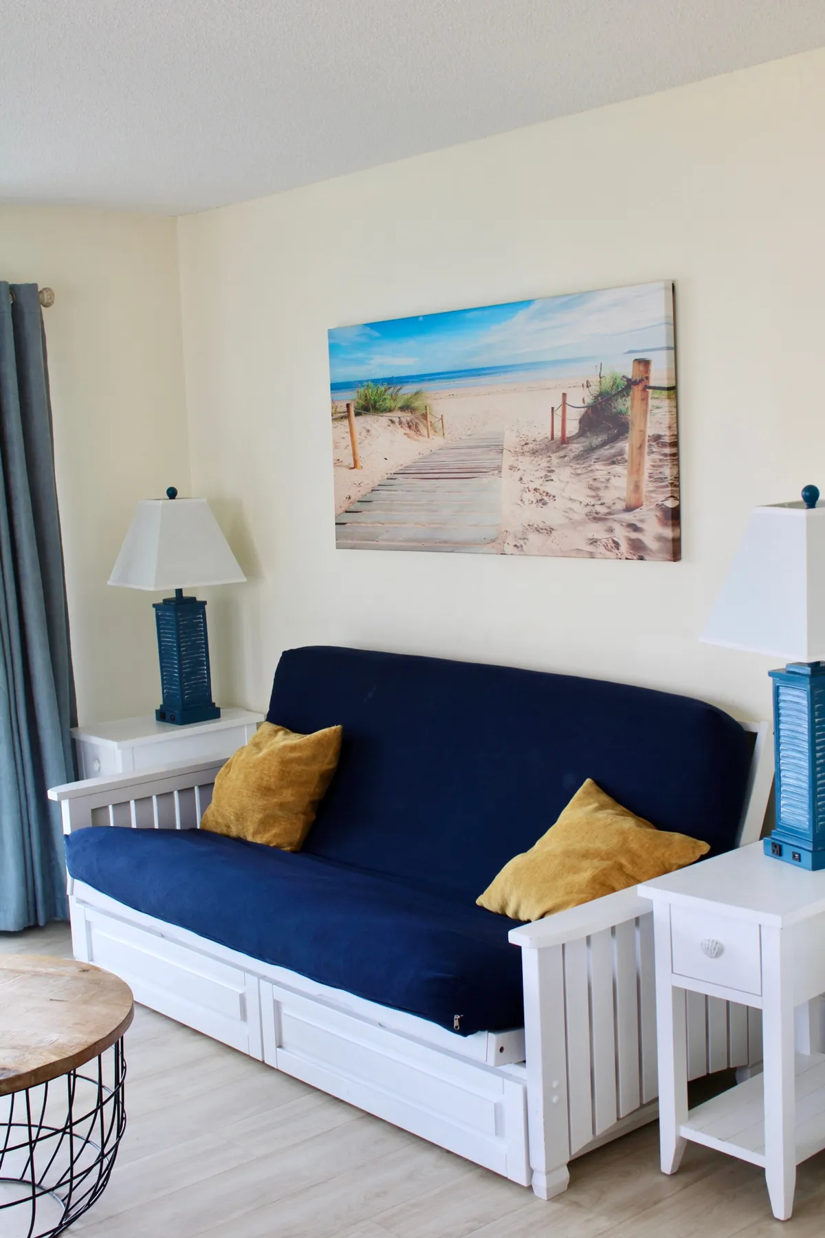 A futon couch is the focal point in this room, accompanied by beach-themed artwork and a soft table lamp. The mood is cozy and beachy, suggesting options for socializing, lounging, or using the futon as a sleeper sofa.