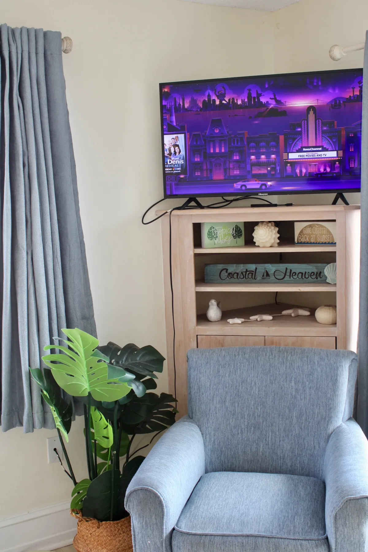 A cozy corner of the living area, complete with a comfortable armchair, a television displaying colorful content, a decorative shelf with coastal-themed items, and a large leafy plant. The setting is casual and welcoming, suggesting both relaxation and beachside charm.