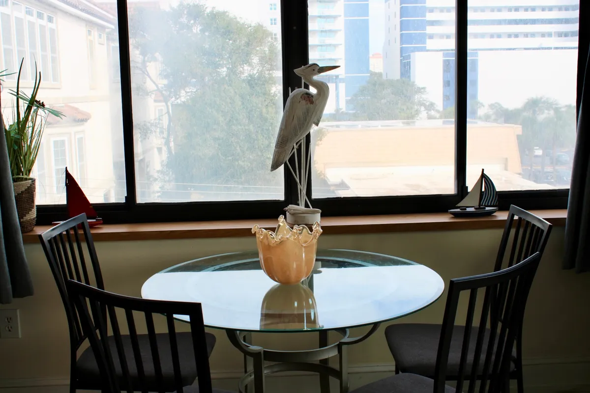 A serene dining nook featuring a glass-topped table set by a window, accompanied by a decorative heron statue and an ornamental bowl. Morning light streams in, hinting at beautiful sunrise views over the ocean, while the intimate setup invites cozy conversations or quiet moments shared between couples.