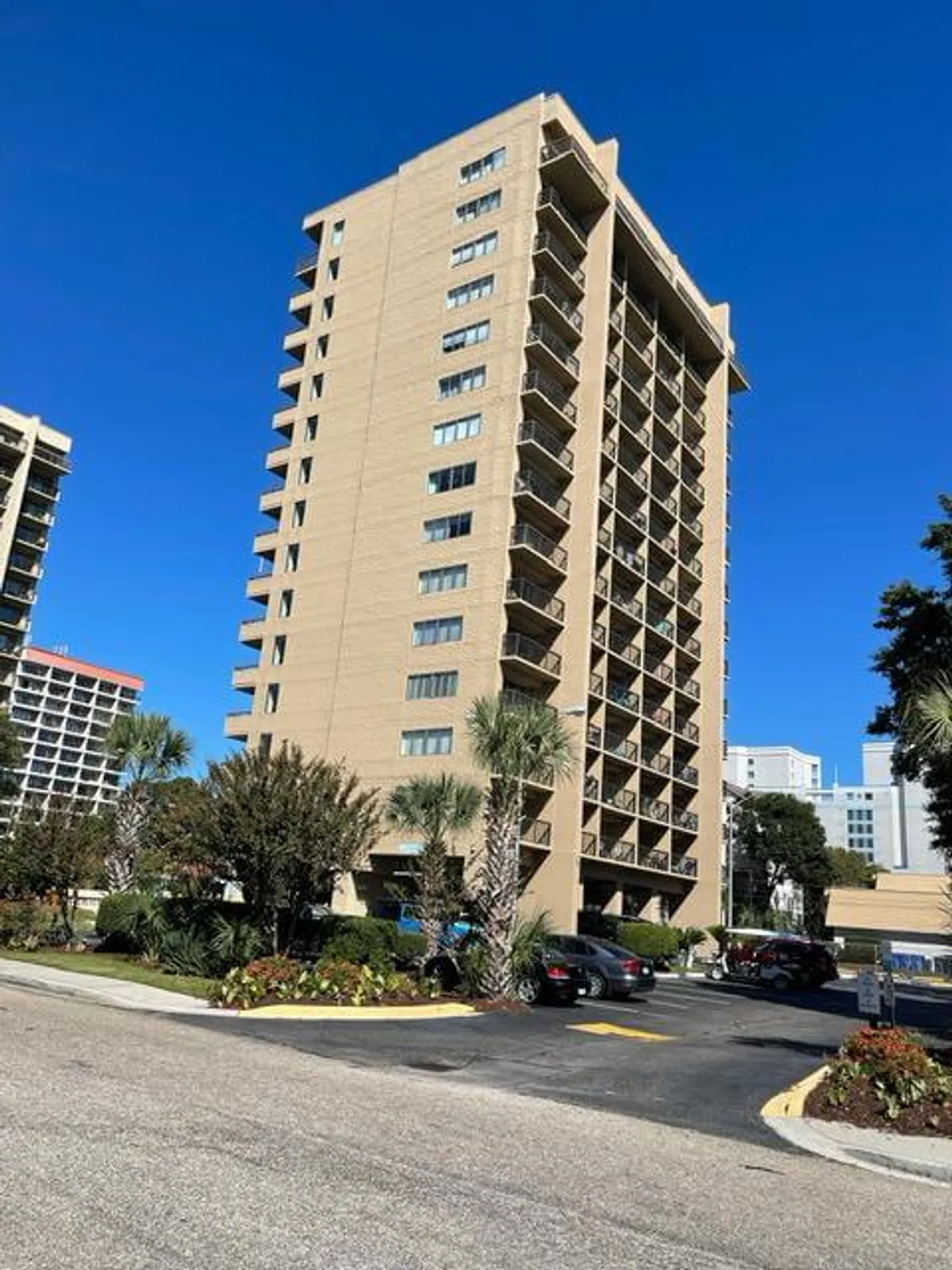 A welcoming exterior shot of a tall residential building surrounded by landscaped gardens and palm trees, with balconies overlooking the grounds. The communal outdoor spaces and inviting environment suggest opportunities for family gatherings and relaxation after a day at the beach.