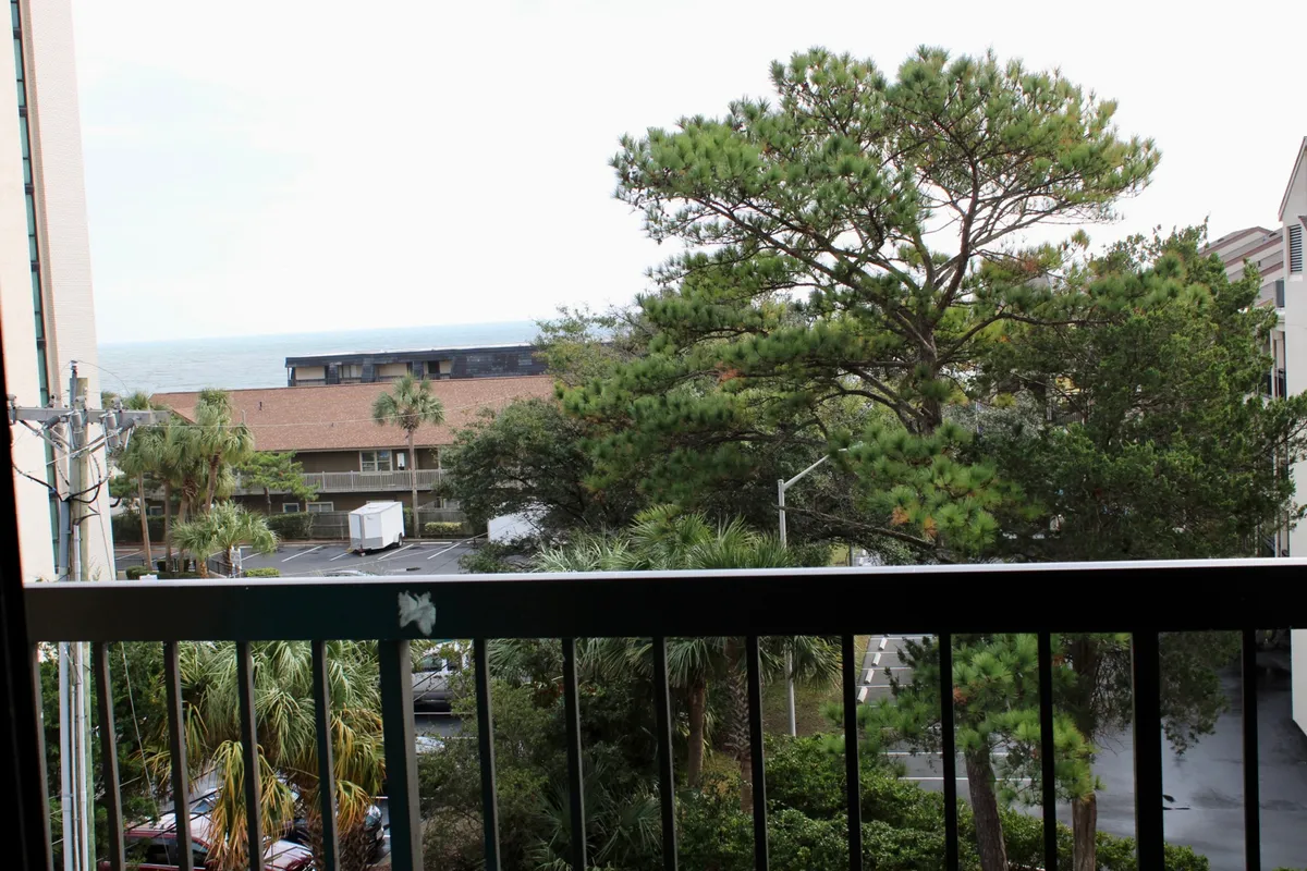 This image presents a tranquil ocean view from a balcony, with tall pine trees and lush greenery framing the sparkling Atlantic. The perspective suggests the viewer is standing on the private balcony, enjoying the expansive coastal scenery and the inviting outdoor atmosphere.